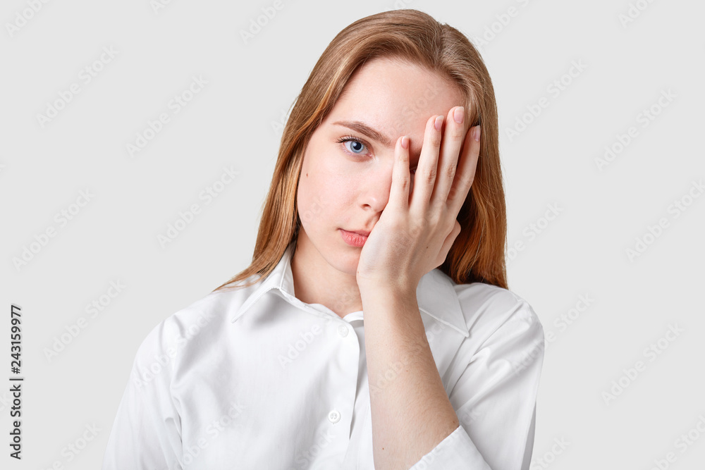 Dissatisfied hopeless blue eyed woman covers face with palm, has long hair, wears elegant shirt, looks stresfully at camera, models against white background. Negative human facial expressions Dissatisfied hopeless blue eyed woman covers face with palm, has long hair, wears elegant shirt, looks stresfully at camera, models against white background. Negative human facial expressions