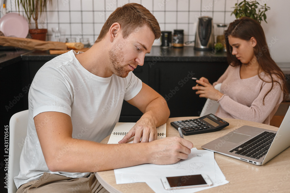 Fototapeta premium Portrait of handsome adult male studying at home, sitting at table in front of portable computer, making notes in copybook, having online marketing courses, cute woman using cell phone in background