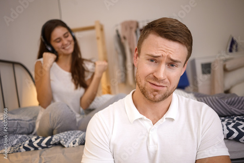 Close up image of frustrated young bearded guy frowning, having painful look, can't sleep because his wife listening to music in background using headphones and dancing. Selective focus on man