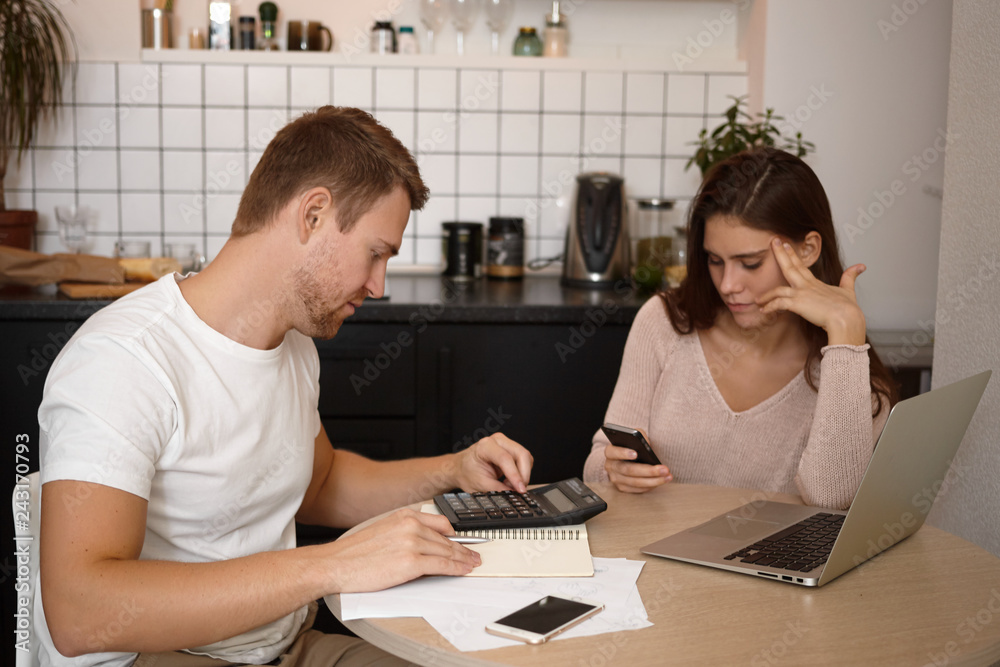 Unshaven Caucasian male calculating family expenses, paying domestic utility bills online using calculator and laptop, his young wife sitting at table with him and typing text message via smart phone