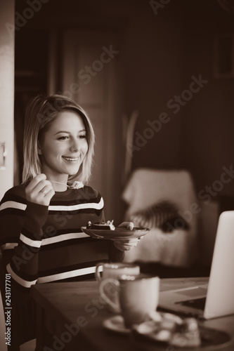 Young lady in sweater sitting at the tablebreakfast time with cup of coffee and cake and looking in to notebook computer. Image in sepia color style photo