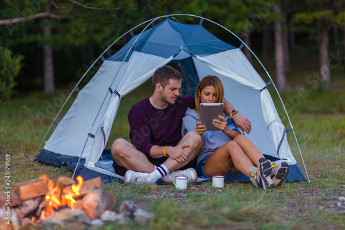 Young couple drinking coffee, enjoying the mountain view and checking the tablet with bonfire in the sunset