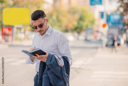 A modern young businessman in white shirt is standing in the city and looking serious while reading a book.