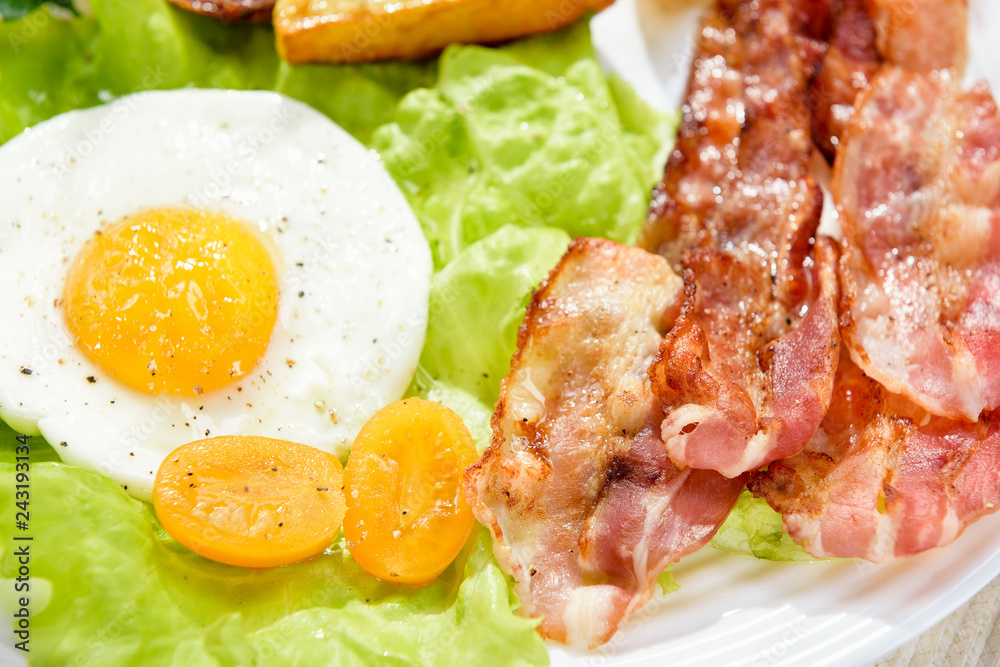Egg with bacon. Roast potatoes. French fries and egg with bacon on top in a white bowl, on a large table with greens. Egg with bacon. Roast potatoes. French fries and egg with bacon on top in a white bowl, on a large table with greens.