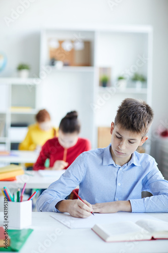 Diligent schoolboy in blue shirt making notes or drawing by desk with two girls on background