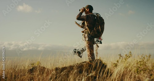 Orbit shot of majestic bowhunter on grassy knoll with his binoculars raised photo