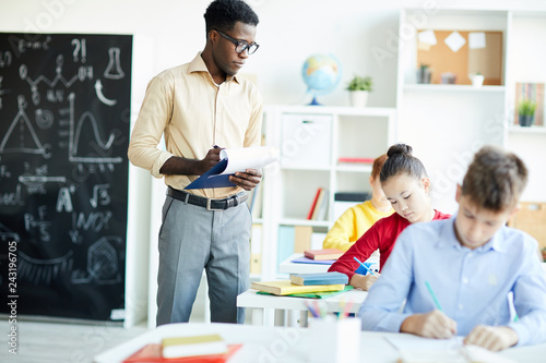 Serious young teacher making notes in his document while standing by one of schoolkids at lesson