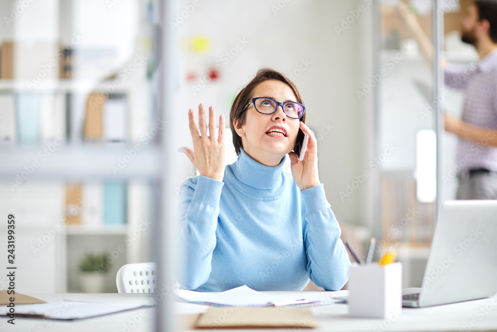 Annoyed office manager explaoinign something to client on the phone while sitting by desk in office Annoyed office manager explaoinign something to client on the phone while sitting by desk in office