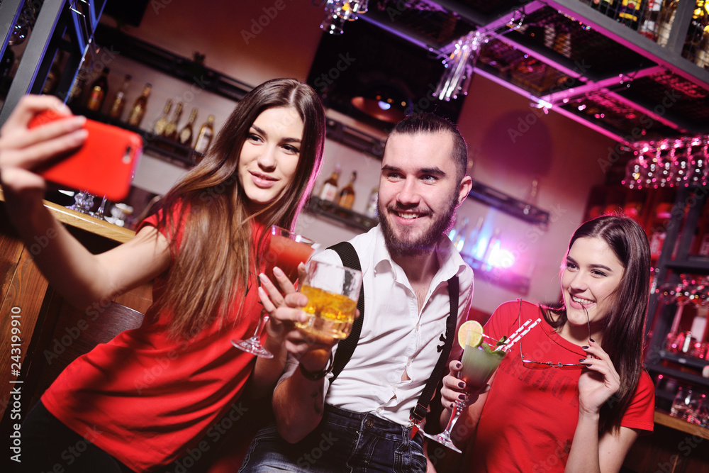 three friends-a young guy and two cute girls holding alcoholic cocktails make a selfie on the background of a bar or a nightclub