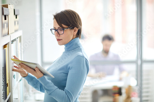 Confident accountant with tablet standing by shelf with archive folders and looking what data should be checked or found