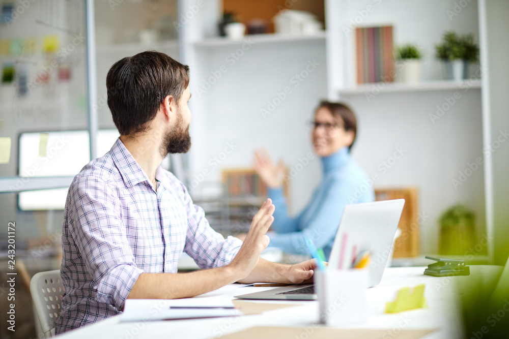 Two office managers sitting by desks and waving by hands to each other while saying something during work Two office managers sitting by desks and waving by hands to each other while saying something during work