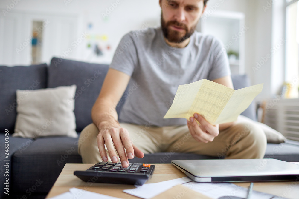 Contemporary young man holding payment bill while pressing calculator buttons and counting monthly budget