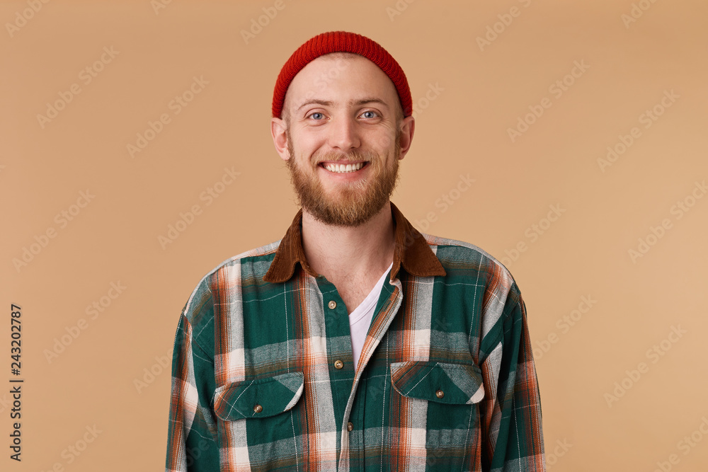 Attractive bearded man in red hat with broad smile showing healthy teeth. A close up portrait of young happy cheerful young man in checkered shirt in front of beige background.