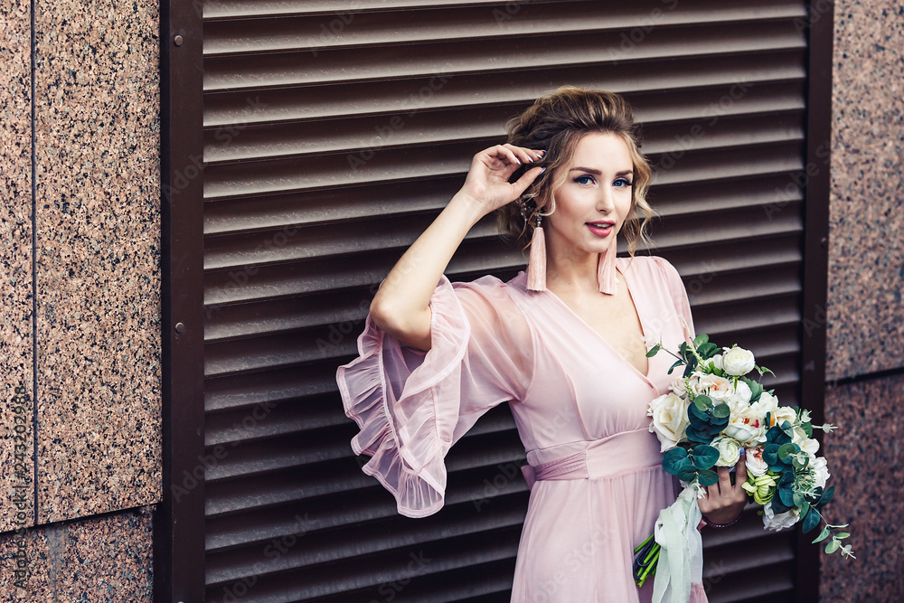Fototapeta premium attractive young girl in a short dress with a bouquet of flowers posing near the granite wall with rolling shutters.
