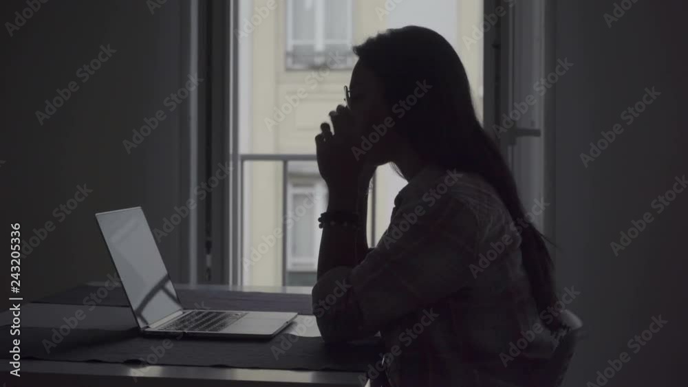 Silhouette of tired young woman with headache working with laptop. Side view of tired businesswoman working on project at home. Overwork concept