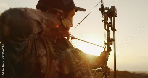 Camera tilts up to bowhunter drawing his bow and aiming into the sunset photo