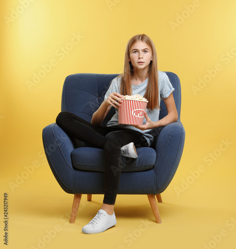Emotional teenage girl with popcorn sitting in armchair during cinema show on color background