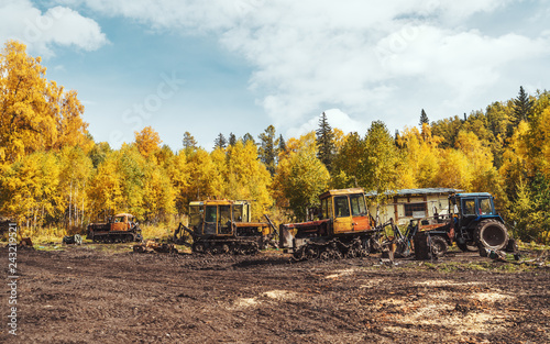 Wide-angle shot of heavy equipment used for logging on a muddy ground filled with tire tracks overlooking a fall coloured forest of birch trees and a partly clouded sky