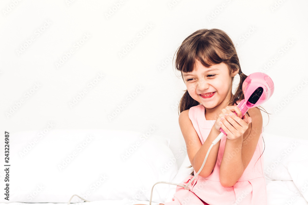 Smiling little girl dries the hair dryer sitting at home on the bed Smiling little girl dries the hair dryer sitting at home on the bed