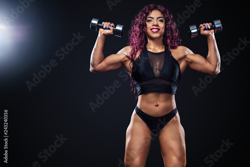 Sporty fit woman, athlete, egyptian and muslim with dumbbells makes fitness exercising on black background with lights.