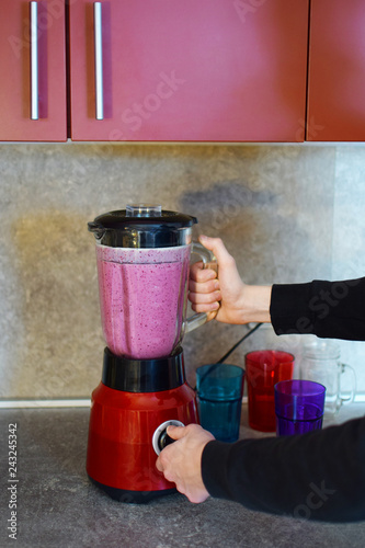 Man hands preparing pink fresh juicy smoothie in red electric blender with glass container, colorful glasses ready for pouring beverage on grey table. Kitchen equipment and healthy food concept.