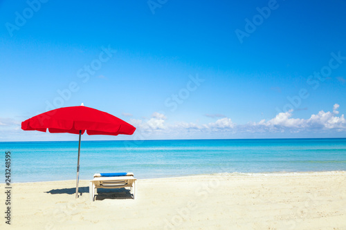 A sun lounger under red umbrella on the sandy beach by the sea and sky. Vacation background. Idyllic beach landscape.
