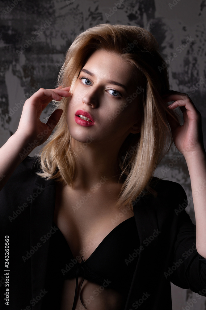 Attractive blonde model with perfect skin wearing black jacket and bra posing at studio with shadows Attractive blonde model with perfect skin wearing black jacket and bra posing at studio with shadows