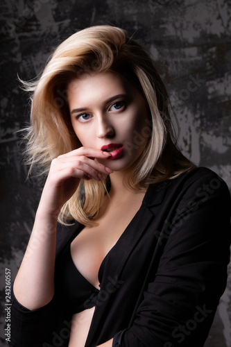 Luxury blonde girl with perfect skin wearing black jacket and bra posing at studio with shadows