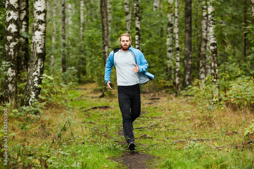 Young active man running down forest path surrounded by green grass and trees on summer weekend Young active man running down forest path surrounded by green grass and trees on summer weekend