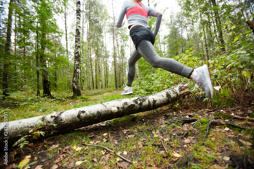 Young active woman in leggins, shorts, sneakers and pullover running over birch log while training in the forest