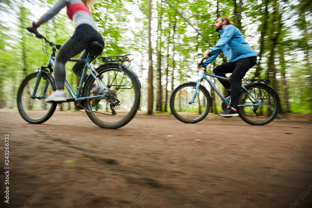 Young active couple riding their bicycles along road surrounded by group of blurry green trees