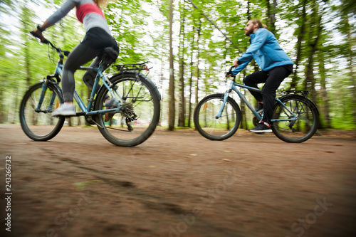 Young active couple riding their bicycles along road surrounded by group of blurry green trees
