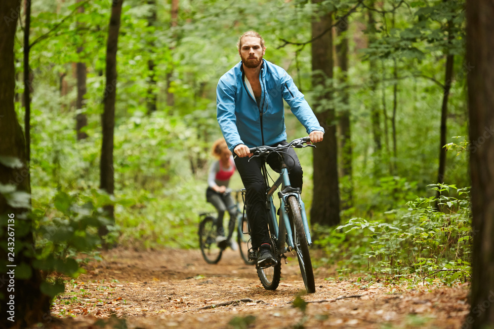 Active guy in sportswear cycling along forest path on summer day with his girlfriend moving after him Active guy in sportswear cycling along forest path on summer day with his girlfriend moving after him