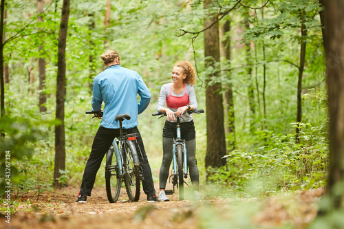 Young female cyclist in activewear talking to her boyfriend during chill in the forest on summer day