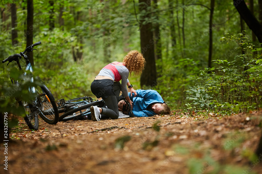 Young sportswoman leaning over male cyclist with broken leg on forest path and trying to understand what to do Young sportswoman leaning over male cyclist with broken leg on forest path and trying to understand what to do