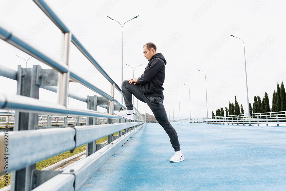 Side view - a handsome slender young man in a gray tracksuit kneads the joints and ligaments of the legs to a street workout standing on the bridge. Copyspace