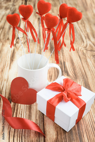 White gift box with red riibon bow, white mug with red hearts on stick, heart card and roses flowers from satin ribbon on wooden background.  photo