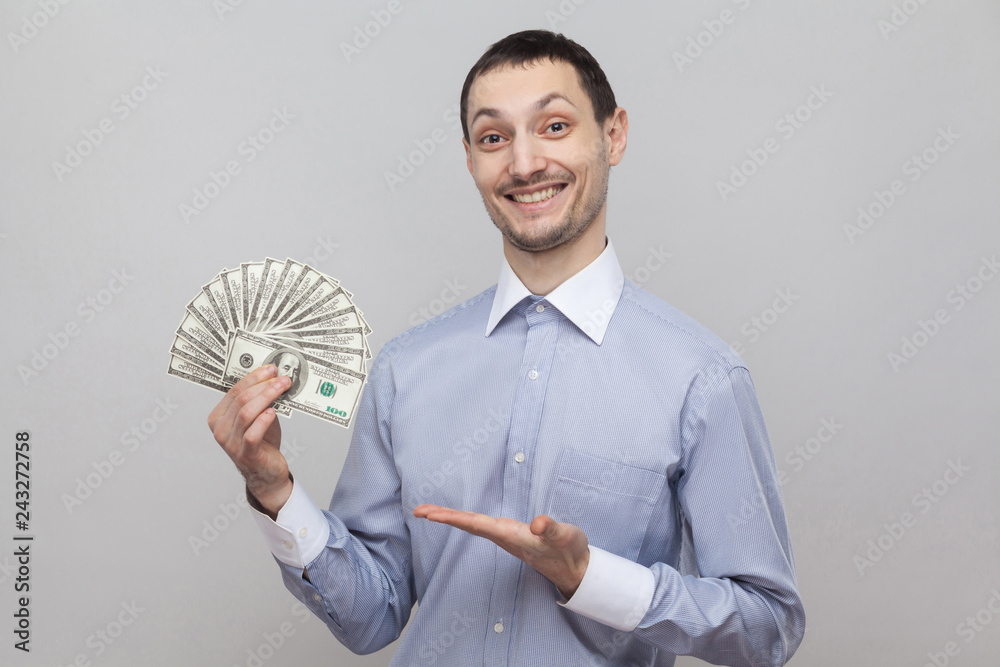 Φωτογραφία Portrait of satisfied attractive young adult businessman in blue shirt standing, giving and pointing with finger fan of money and toothy smile