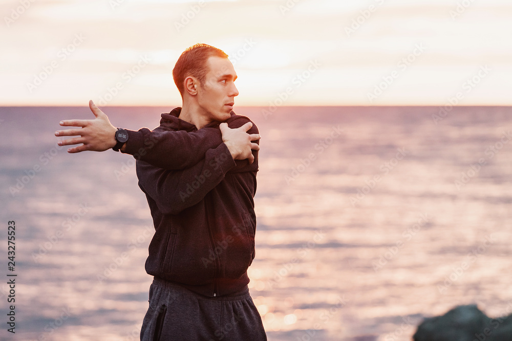 Side view of a slim tall guy athlete doing a warm-up on a rocky seashore against the backdrop of the sunset. Concept of sports training after a hard day. Copyspace Side view of a slim tall guy athlete doing a warm-up on a rocky seashore against the backdrop of the sunset. Concept of sports training after a hard day. Copyspace