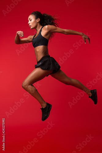 Portrait of energetic african american woman in black sportswear running, isolated over red background