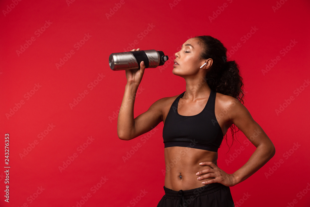 Image of healthy african american woman in black sportswear holding water bottle, isolated over red background