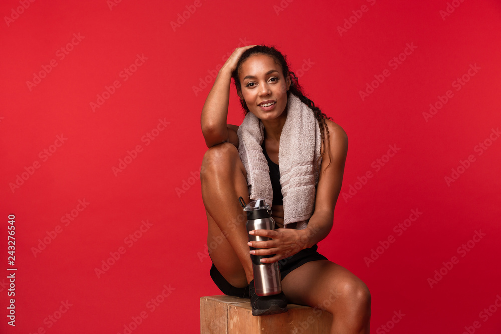 Obraz premium Portrait of joyous african american woman in black sportswear sitting on box with towel and water bottle, isolated over red background