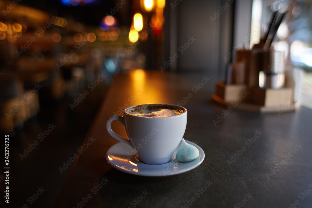 Obraz premium Cup of americano coffee in a white cup with a marshmallow on a saucer, shallow focus, dark background, bulb lights of caffeine
