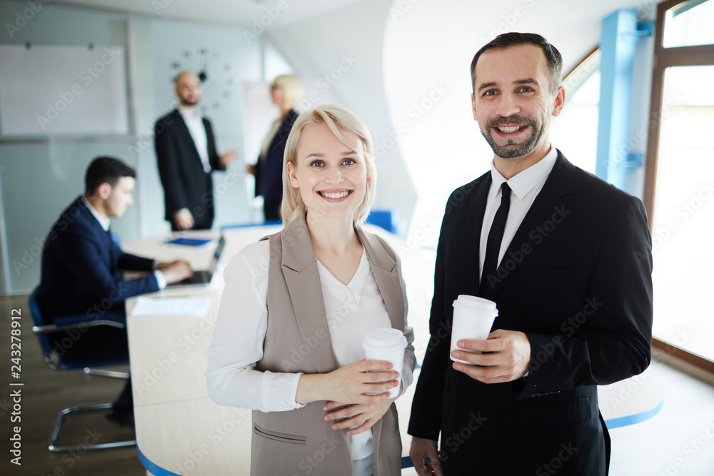 Two elegant cheerful colleagues with glasses of coffee looking at you in boardroom at break Two elegant cheerful colleagues with glasses of coffee looking at you in boardroom at break