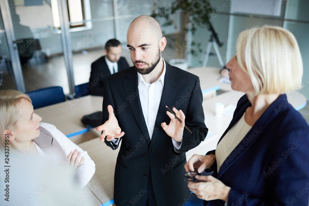 Young confident businessman talking to his female colleagues during discussion of financial news or data Young confident businessman talking to his female colleagues during discussion of financial news or data