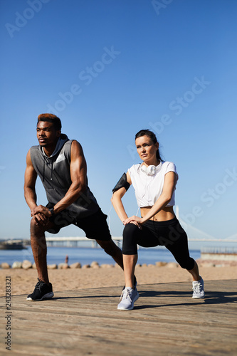 Serious concentrated young interracial couple in sportswear leaning on knees while stretching legs, they practicing warmup outdoors