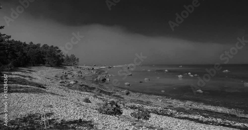 Black and white time lapse of clouds moving over a still sea on a rocky shore with trees