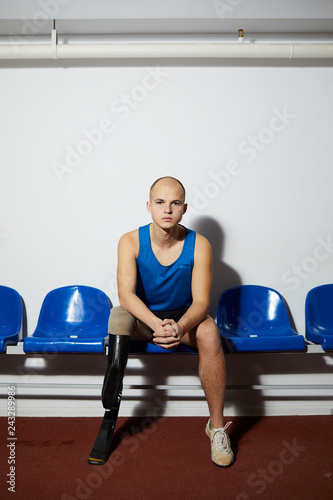 Disable young sportsman with handicap sitting on blue plastic chair along wall before competition photo