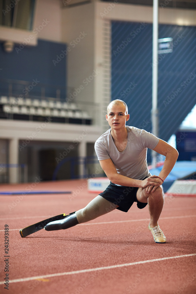 Fototapeta premium Young man in activewear doing stretching exercise for legs on racetrack on stadium while preparing for paralympic games
