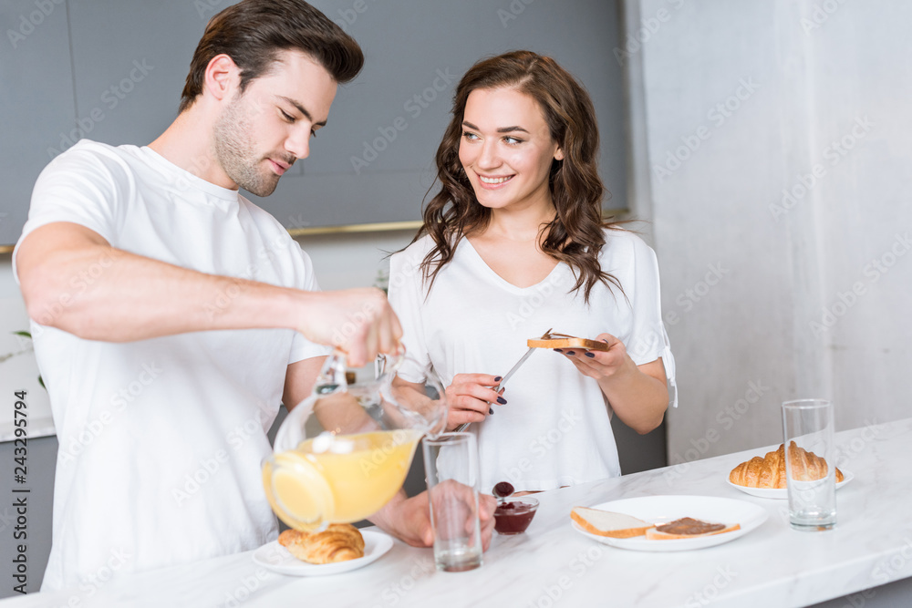 girlfriend looking at handsome man holding jug with orange juice in kitchen